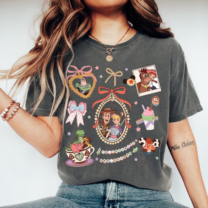 May include: Dark grey t-shirt with colourful cartoon graphics of Toy Story characters. The design includes images of Woody, Buzz Lightyear, and other characters, along with bows and stars.