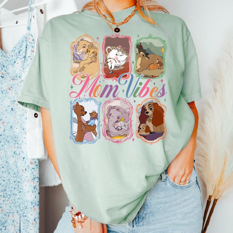 May include: A light green t-shirt featuring six framed cartoon images of mothers and their babies. The words "Mom Vibes" are written in a colorful, cursive font across the center of the shirt. The shirt has short sleeves and a crew neck.