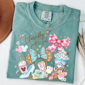 May include: A teal Comfort Colors t-shirt with the phrase "Literally just a girl" and various cartoon graphics, including a high heel, popcorn, and a bow. A silver necklace is draped across the shirt.