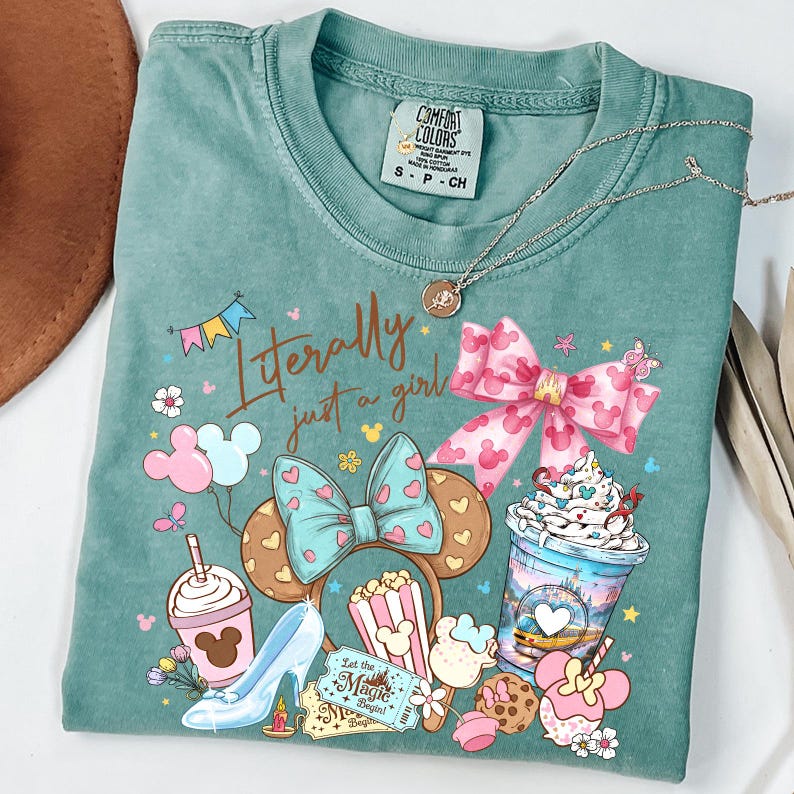 May include: A teal Comfort Colors t-shirt with the phrase "Literally just a girl" and various cartoon graphics, including a high heel, popcorn, and a bow. A silver necklace is draped across the shirt.