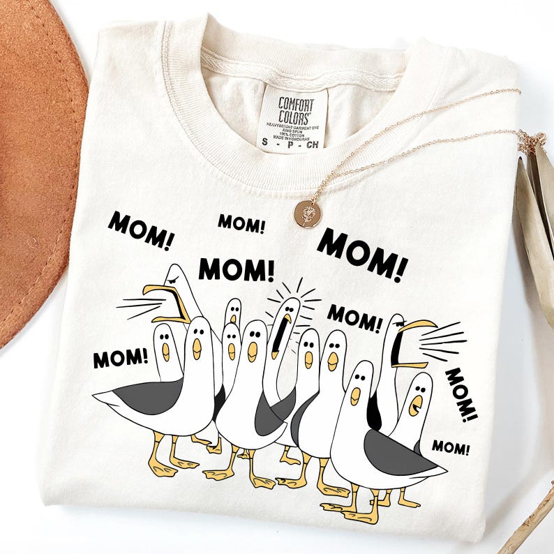 May include: Cream-colored t-shirt with a graphic of cartoon seagulls and the word "MOM!" repeated. The seagulls are white with gray wings and yellow feet. A gold necklace with a pendant is draped on the shirt.