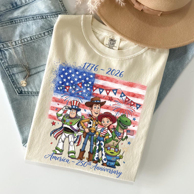 May include: Cream-colored t-shirt featuring a graphic of the American flag and characters from the Toy Story movie. The shirt has the text "1776-2026" and "America - 250th Anniversary". The shirt is paired with blue jeans and a tan hat.