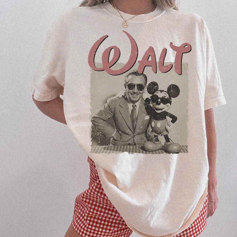May include: Cream-colored t-shirt featuring a sepia-toned image of Walt Disney with Mickey Mouse. The word "Walt" is printed above the image in a pink, cursive font. The shirt has short sleeves and a relaxed fit.