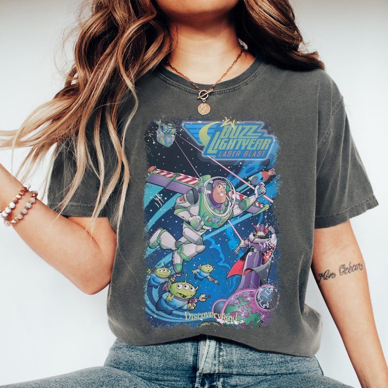 K&ouml;nnte beinhalten: Dunkelgraues T-Shirt mit einer farbenfrohen Grafik von Buzz Lightyear im Weltraum, mit dem Text "Buzz Lightyear Laser Blast" und "Discoveryland". Das Design umfasst Aliens und einen lila Schurken.