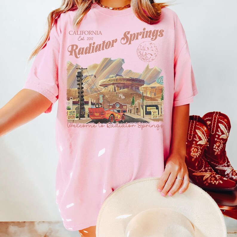 May include: Pink t-shirt with a graphic of Radiator Springs, California, featuring a car and buildings. Text includes "CALIFORNIA Est. 2012" and "Welcome to Radiator Springs". The shirt is paired with red cowboy boots.