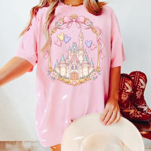 May include: Pink t-shirt featuring a castle illustration within a decorative frame with a bow and flowers. The castle has multiple turrets and is surrounded by balloons. The shirt is paired with red cowboy boots.