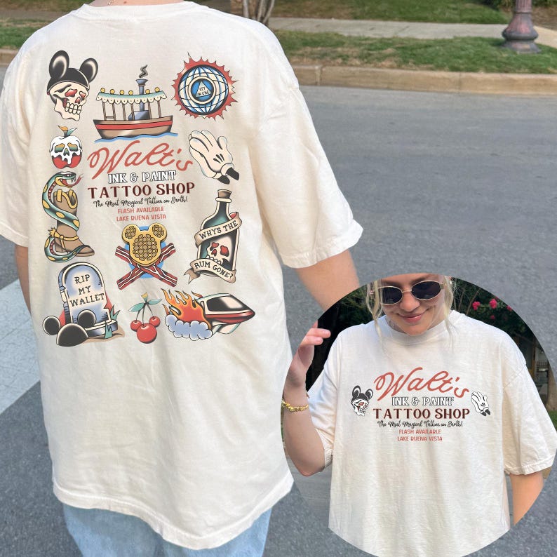May include: Cream-colored t-shirt featuring a tattoo-style design with the text "Walt's Ink & Paint Tattoo Shop." The back of the shirt displays various tattoo-inspired illustrations, including a skull, boat, and snake.