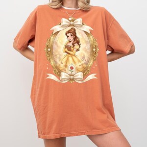 Op de afbeelding: Oranje T-shirt met een ingelijste illustratie van een vrouw in een gele jurk die een boek vasthoudt. De gouden lijst heeft witte strikken en een rode roos. De vrouw heeft bruin haar en glimlacht.