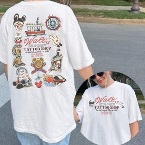 May include: White t-shirt featuring colorful tattoo-style illustrations and text. The design includes a skull with Mickey Mouse ears, a boat, a hand, a bottle, a snake, a tombstone, cherries, and a spaceship. The text reads "Walt's Ink & Paint Tattoo Shop".