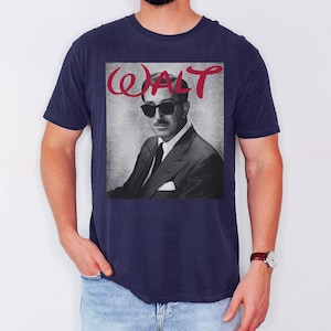 May include: Navy blue t-shirt featuring a black and white image of a man in a suit and sunglasses. The word "WALT" is printed in red cursive above the image. The shirt is a classic crew neck style.