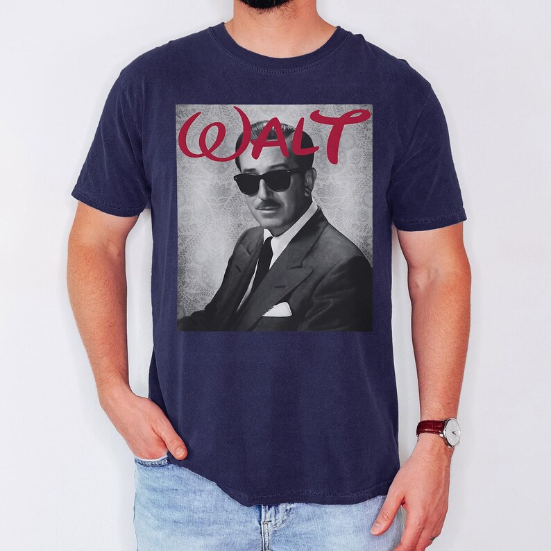 May include: Navy blue t-shirt featuring a black and white image of a man in a suit and sunglasses. The word "WALT" is printed in red cursive above the image. The shirt is a classic crew neck style.