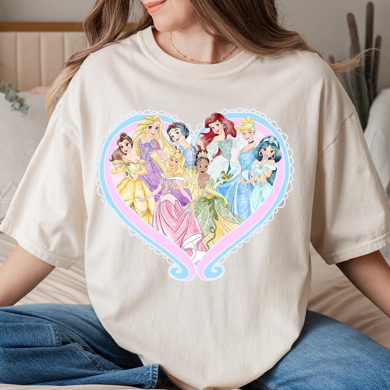 May include: Cream-colored t-shirt featuring a heart-shaped design with illustrations of various princesses in colorful gowns. The heart is outlined in pink and blue with a decorative border. The shirt is made of a soft material.