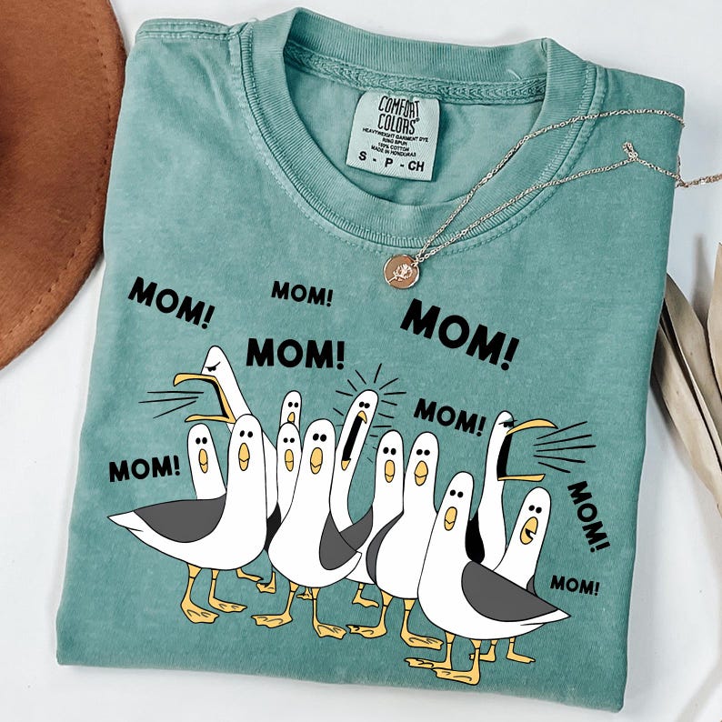 May include: A teal Comfort Colors t-shirt featuring a graphic of cartoon seagulls with the word "MOM!" repeated around them. A silver necklace with a pendant is draped on the shirt.