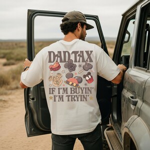 May include: Cream-colored t-shirt with the text "DAD TAX" and the phrase "IF I'M BUYIN' I'M TRYIN'" in brown and gold. The shirt features cartoon illustrations of treats and Mickey Mouse.