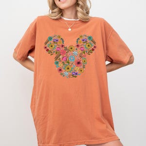 K&ouml;nnte beinhalten: Orangefarbenes T-Shirt mit einem floralen Mickey-Mouse-Design. Das Design besteht aus verschiedenen bunten Blumen, darunter Rosa, Gelb, Blau und Lila, die in Form des Mickey-Mouse-Kopfes angeordnet sind. Das Shirt hat kurze &Auml;rmel und eine lockere Passform.