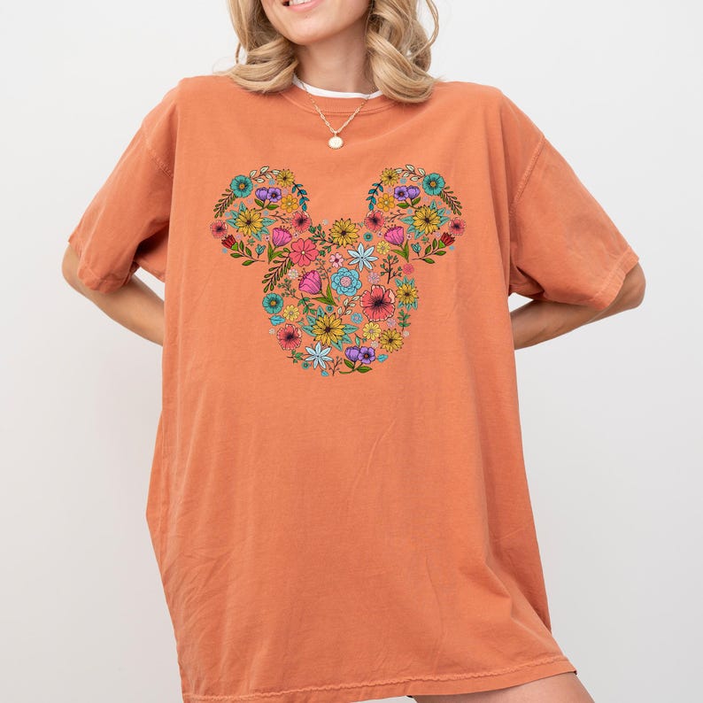 K&ouml;nnte beinhalten: Orangefarbenes T-Shirt mit einem floralen Mickey-Mouse-Design. Das Design besteht aus verschiedenen bunten Blumen, darunter Rosa, Gelb, Blau und Lila, die in Form des Mickey-Mouse-Kopfes angeordnet sind. Das Shirt hat kurze &Auml;rmel und eine lockere Passform.