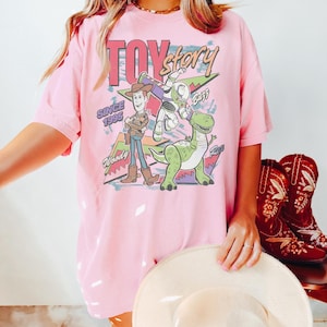 May include: Pink t-shirt featuring a graphic of Woody, Buzz Lightyear, and Rex from Toy Story. The design includes the text "TOY Story" and "Since 1995". The shirt has short sleeves and a relaxed fit.