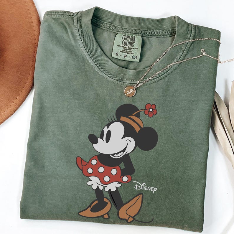 Może przedstawiać: Oliwkowo-zielona koszulka Comfort Colors z grafiką Myszki Minnie. Myszka Minnie ma na sobie czerwoną sukienkę w białe grochy, brązowe buty i kwiat we włosach. Złoty naszyjnik z wisiorkiem spoczywa na koszulce. Widoczne jest logo Disneya.