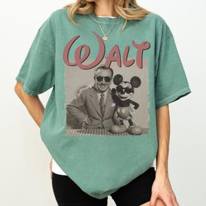 May include: A seafoam green t-shirt featuring a vintage-style graphic. The graphic includes the name "Walt" in a cursive font, along with a sepia-toned image of Walt Disney and Mickey Mouse wearing sunglasses. The shirt has a relaxed fit.