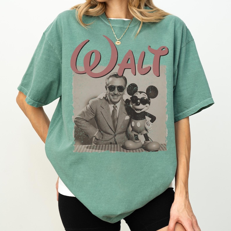 May include: A seafoam green t-shirt featuring a vintage-style graphic. The graphic includes the name "Walt" in a cursive font, along with a sepia-toned image of Walt Disney and Mickey Mouse wearing sunglasses. The shirt has a relaxed fit.