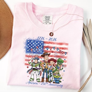 May include: A light pink t-shirt featuring a graphic of the American flag with fireworks and cartoon characters. The text "1776-2026" and "America - 250th Anniversary" are printed on the shirt. A rose gold necklace is draped over the shirt.