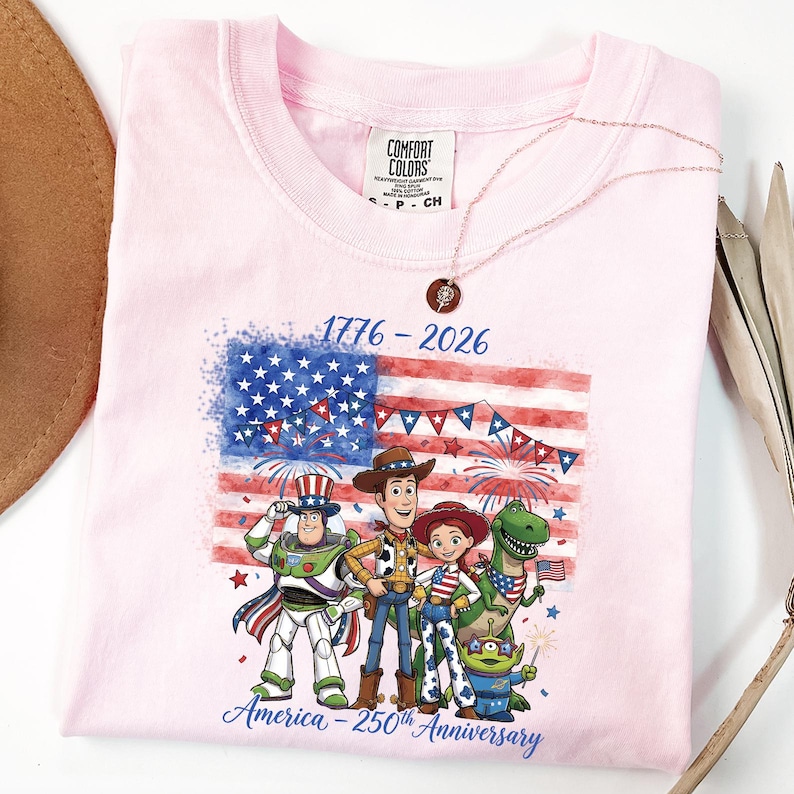 May include: A light pink t-shirt featuring a graphic of the American flag with fireworks and cartoon characters. The text "1776-2026" and "America - 250th Anniversary" are printed on the shirt. A rose gold necklace is draped over the shirt.