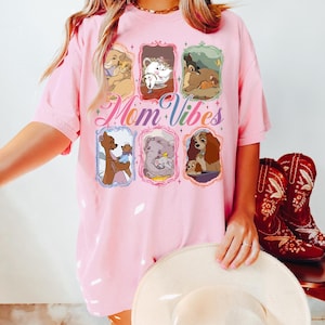 May include: Pink t-shirt featuring cartoon images of Disney characters in ornate frames. The words "Mom Vibes" are written in a script font. A woman is wearing the shirt and holding a white hat. Red cowboy boots are in the background.