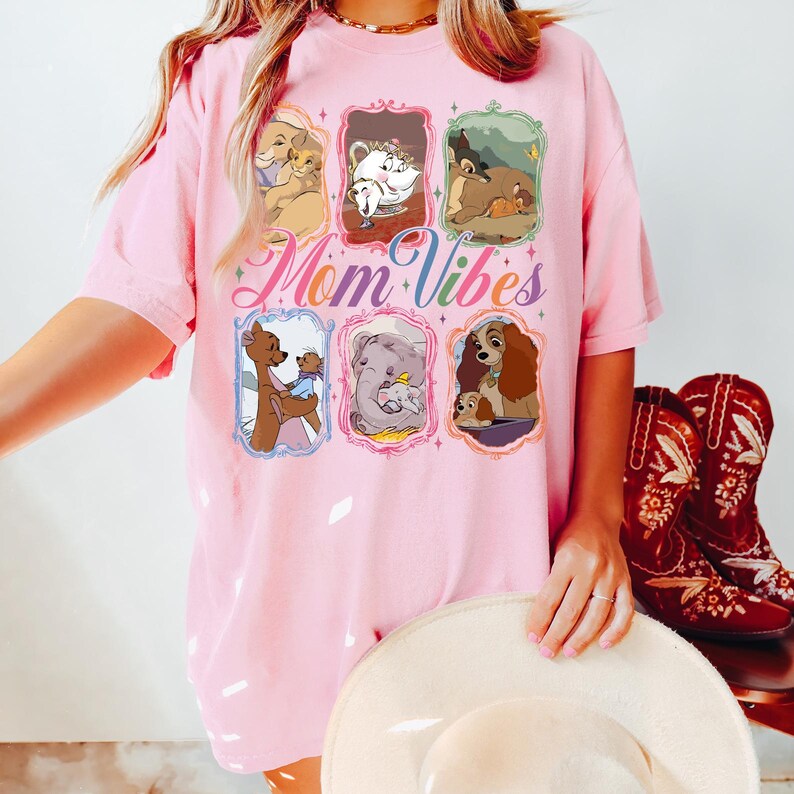 May include: Pink t-shirt featuring cartoon images of Disney characters in ornate frames. The words "Mom Vibes" are written in a script font. A woman is wearing the shirt and holding a white hat. Red cowboy boots are in the background.