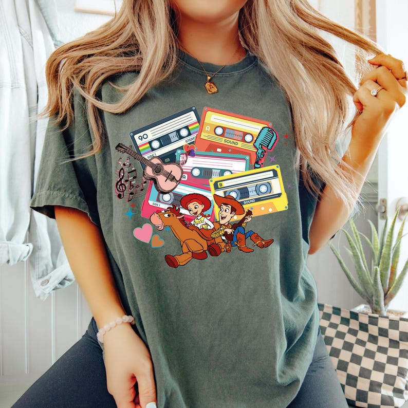 Puede incluir: Camiseta verde oscuro con un gr&aacute;fico colorido de cintas de cassette, una guitarra, notas musicales y personajes de dibujos animados. El dise&ntilde;o incluye un vaquero y una vaquera montados a caballo. La camiseta es de material suave.