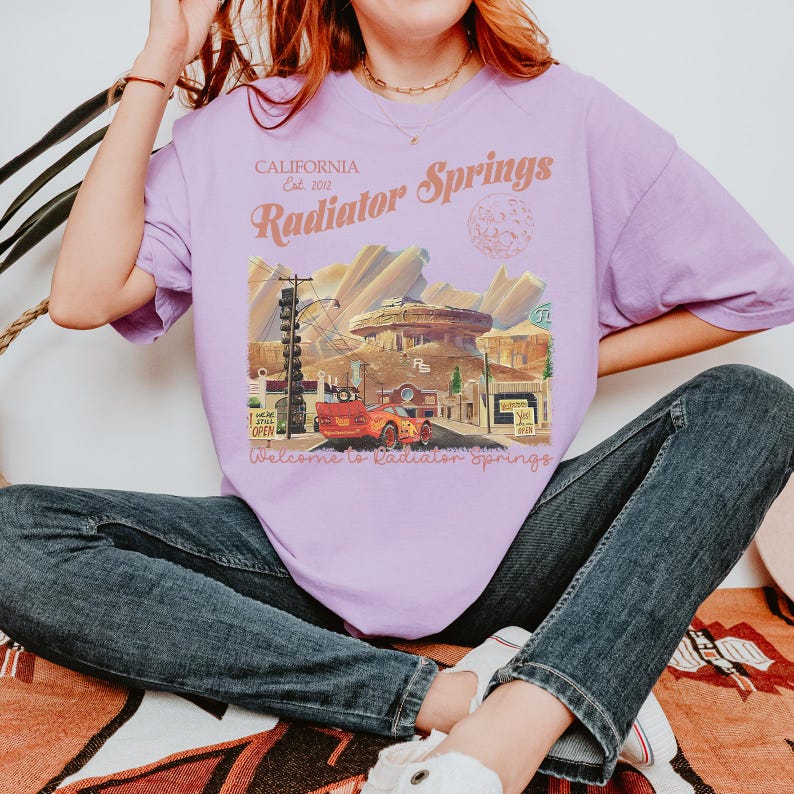May include: A light purple t-shirt featuring a graphic of Radiator Springs from the movie Cars. The graphic includes the text "CALIFORNIA Est. 2012 Radiator Springs" and "Welcome to Radiator Springs". The shirt is a relaxed fit.