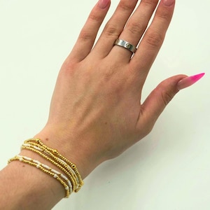 May include: A silver ring with gold accents and a beaded bracelet set. The bracelets feature gold and white beads. The nails are painted with a pink ombre design.