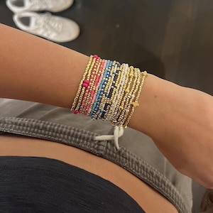 May include: A stack of multiple beaded bracelets featuring a variety of colors including gold, pink, blue, white, and black. The bracelets are composed of small round beads, with some strands incorporating gold-toned cross charms.