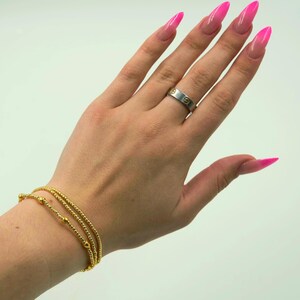 May include: A hand wearing a silver ring with gold accents and a gold bracelet with three strands of small beads. The nails are long and painted a bright pink.