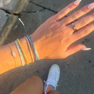 May include: A collection of four bracelets and one ring. The bracelets include a gold chain with an oval charm, a white and blue beaded strand, a gold beaded strand, and a light blue cord. The ring is a gold band with screw head motifs.