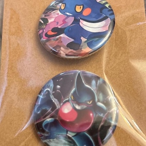 May include: Two round pinback buttons featuring cartoon characters. One button has a blue character with orange cheeks and yellow eyes. The other button has a dark blue character with a red chest and a pointed head. Both buttons have colorful backgrounds.