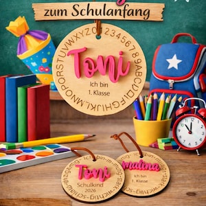Op de afbeelding: Drie ronde houten labels met leren koordjes. Een groot label bevat de naam Toni, het alfabet en de tekst Ich bin 1. Klasse. Twee kleinere labels bevatten de namen Toni en Malina met de tekst Ich bin 1. Klasse en Schulkind 2026.