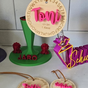 Op de afbeelding: Een verzameling gepersonaliseerde items, waaronder een ronde houten hanger met de naam Toni, een groene standaard met de naam Jaro en twee rode dinosaurusfiguren, een paarse kegelvormige houder en twee kleinere ronde houten hangers.