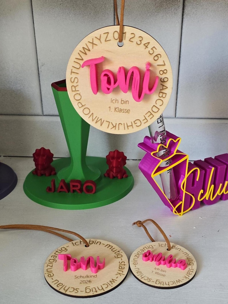 Op de afbeelding: Een verzameling gepersonaliseerde items, waaronder een ronde houten hanger met de naam Toni, een groene standaard met de naam Jaro en twee rode dinosaurusfiguren, een paarse kegelvormige houder en twee kleinere ronde houten hangers.