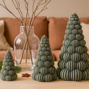 Könnte beinhalten: Drei dekorative Weihnachtsbaum-Skulpturen in Salbeigrün mit geschichtetem Design stehen auf einem Holztisch. Rote Glitzerstern-Ornamente sind um die Bäume herum verstreut. Eine klare Glasvase mit Zweigen und ein Sofa befinden sich im Hintergrund.