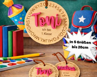 Op de afbeelding: Drie ronde houten hangers met bruine leren koorden. De grootste heeft de naam Toni in roze, het alfabet en de tekst Ich bin 1. Klasse. Twee kleinere hangers hebben de namen Toni en Malina met de tekst Schulkind 2026.
