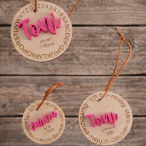 Op de afbeelding: Drie ronde houten ornamenten met bruine leren koorden. E&eacute;n toont de naam Toni in roze met het alfabet en de tekst Ich bin 1. Klasse. De andere tonen de namen Emilia of Toni in roze met de tekst Ich bin mutig-stark-wichtig-schlau.