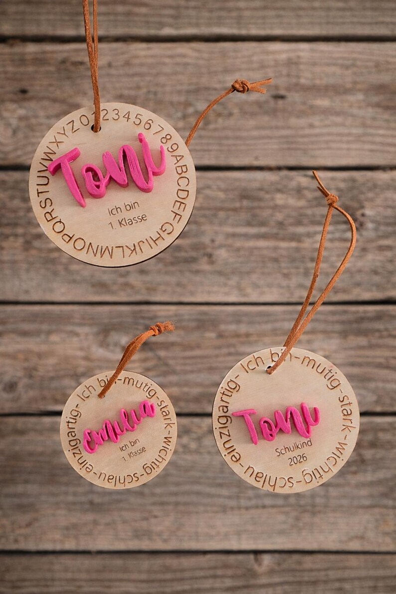 Op de afbeelding: Drie ronde houten ornamenten met bruine leren koorden. E&eacute;n toont de naam Toni in roze met het alfabet en de tekst Ich bin 1. Klasse. De andere tonen de namen Emilia of Toni in roze met de tekst Ich bin mutig-stark-wichtig-schlau.