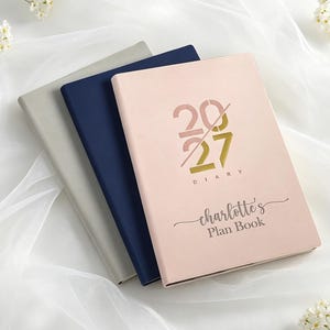 以下が含まれることがあります： 3冊の長方形のプランナーが重なっています。一番上のプランナーはライトピンクで、表紙に「2027 DIARY」と「charlotte's Plan Book」の文字が印刷されています。真ん中のプランナーはダークブルー、一番下のプランナーはライトグレーです。