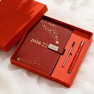 以下が含まれることがあります： 赤いギフトボックスには、お揃いの赤い2026年版デイリープランジャーナルと2本のペンが入っています。ジャーナルには金色のスターバーストのデザインと、銀色の留め金に「William」という名前が記されています。ペンはシルバーとレッドです。