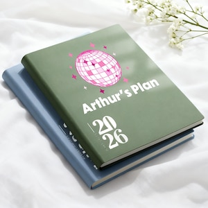 以下が含まれることがあります： 2冊のハードカバープランナー。1冊は水色、もう1冊はオリーブグリーンで、重ねられています。上のプランナーには「Arthur's Plan」と「2026」の文字と、ピンクのディスコボールのグラフィックが描かれています。