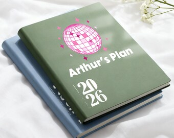 May include: Two hardcover planners, one light blue and one olive green, stacked on top of each other. The top planner has the text "Arthur's Plan" and "2026" with a pink disco ball graphic.