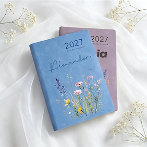 Może przedstawiać: Jasnoniebieski notatnik z harmonogramem na 2027 rok z imieniem Alexander napisanym kursywą i ilustracją polnych kwiat&oacute;w. Częściowo widoczny za nim jest fioletowy notatnik na 2027 rok z nadrukowanym na okładce imieniem Sofia.