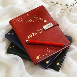 May include: A stack of three leather-bound 2026 daily planners in red, navy, and green. The red planner has gold starburst designs and the text "2026 DAILY PLAN" with a silver nameplate that reads "Daniel". A red pen rests on top.