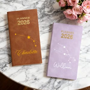 以下が含まれることがあります： 2026年のパーソナライズされたプランナーが2つ。1つは茶色で、金色の文字と星座のデザインがあり、「Charlotte」という名前が筆記体で書かれています。もう1つはラベンダー色で、星座のデザインと「William」という名前が書かれています。