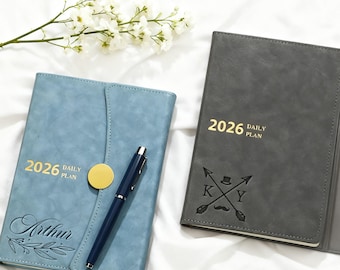 May include: Two 2026 daily plan journals. One is blue with "Arthur" and a floral design, the other is gray with a mustache and arrow design. A blue pen rests on the blue journal. White flowers are in the background.